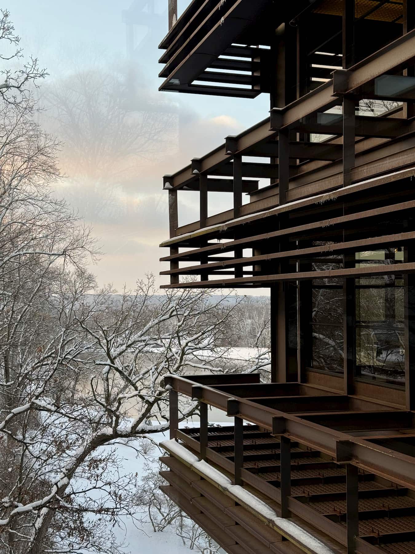 Modern architectural balcony with steel beams and glass windows overlooking a serene, snow-covered landscape with bare trees. The sky is painted with soft hues of blue and orange. The peaceful setting provides a beautiful contrast to the modern building design.