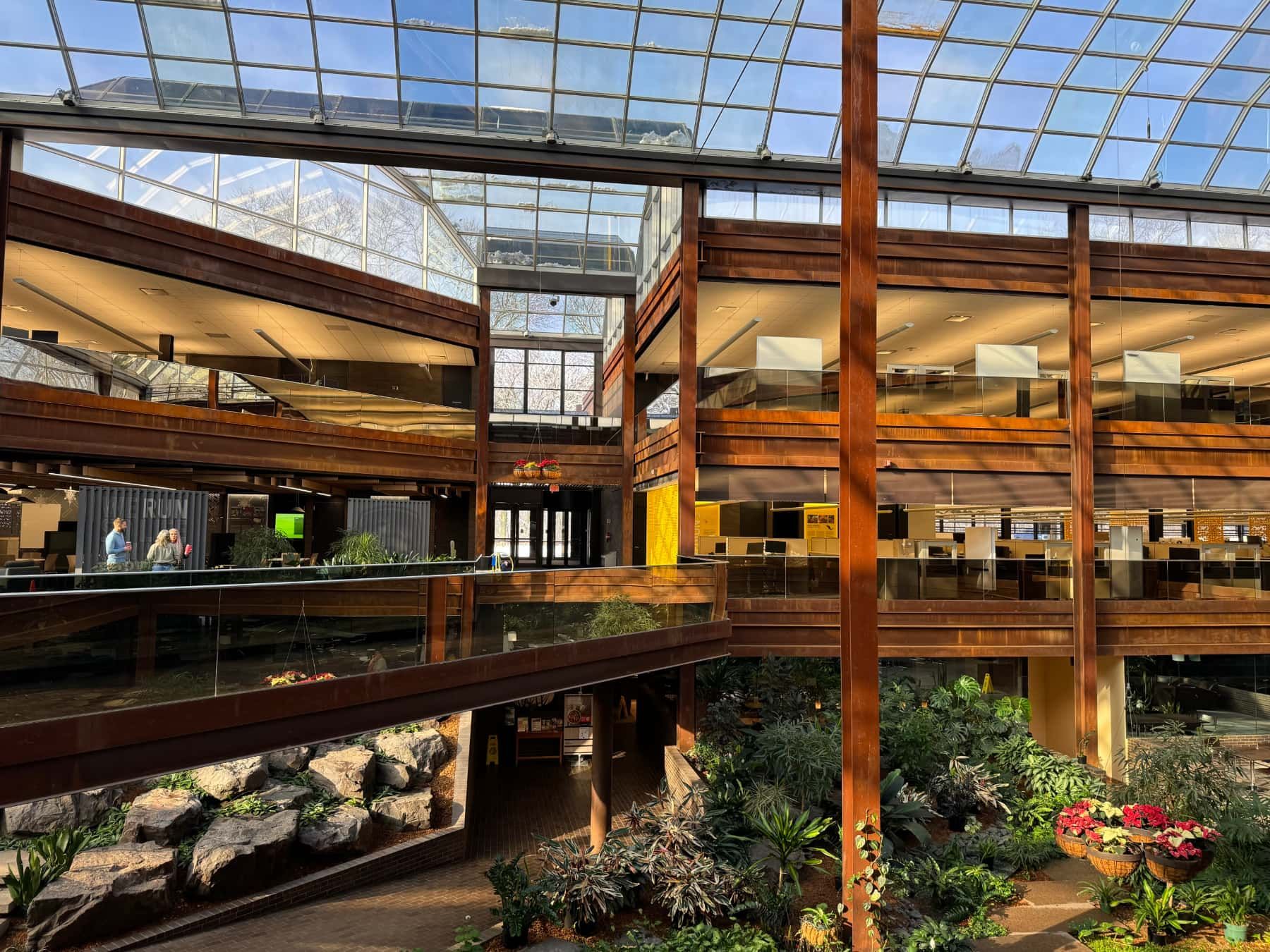 Large indoor space with a garden at the ground level with rust-colored steel balconies on multiple levels. A large glass ceiling allows light to fill the space. The garden has lush green plants and rocks enhancing the natural look. The architecture is modern with clean lines and open spaces allowing for visibility across levels.