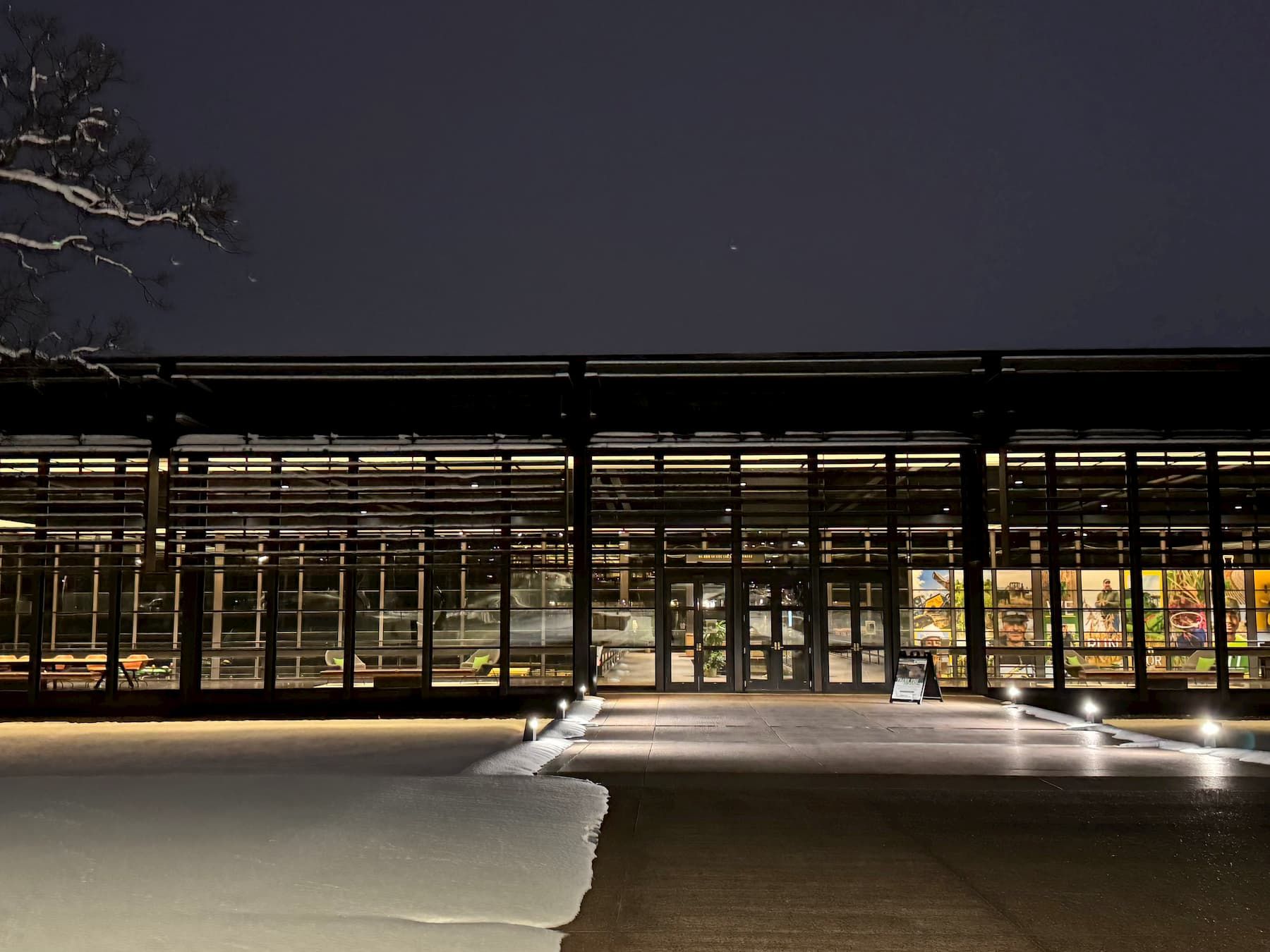 Modern building with large, illuminated glass windows during nighttime. The building is long and rectangular, with visible structural beams. Various colorful displays are inside but not clearly identifiable. The groundis covered in snow, reflecting light from the windows.